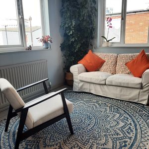 The therapy room. There is a black framed armchair with white cushions facing a white sofa with orange cushions. A large blue and white floral patterned rug is on the floor, and there is a tall plant in the corner of the room. The room is light, with a window on each of the two visible walls.