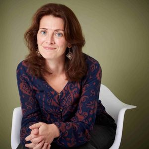 Kate Kingsford. She has reddish brown hair and blue eyes. She is sitting on a white chair, wearing a dark blue and brown patterend top, and has a pair of geometic earrings.